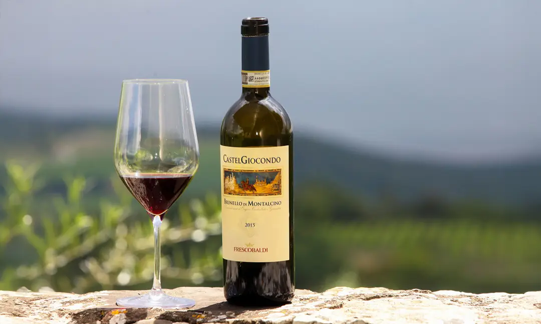 Glass of red wine beside a bottle of Brunello di Montalcino CastelGiocondo 2015, from Frescobaldi winery, with a Tuscan vineyard landscape in the background 