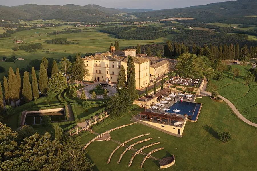 Aerial view of Castello di Casole, a luxurious hotel set in a historic castle surrounded by gardens, cypress trees, and Tuscan hills, with an outdoor pool and elegant facilities.