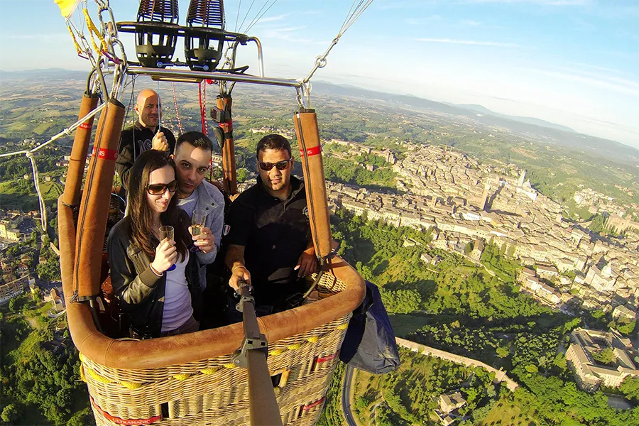 Group of people in a hot air balloon flying over a historic city in Tuscany, Italy, enjoying the view and toasting with glasses during the flight.