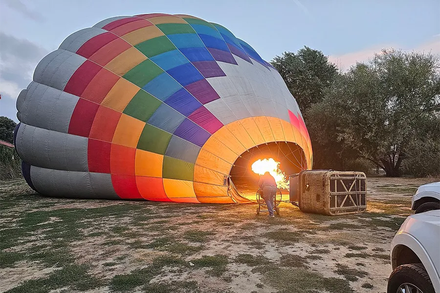 Balão de ar quente multicolorido sendo inflado com chama ao amanhecer, com cesta posicionada ao lado em um campo aberto na Toscana