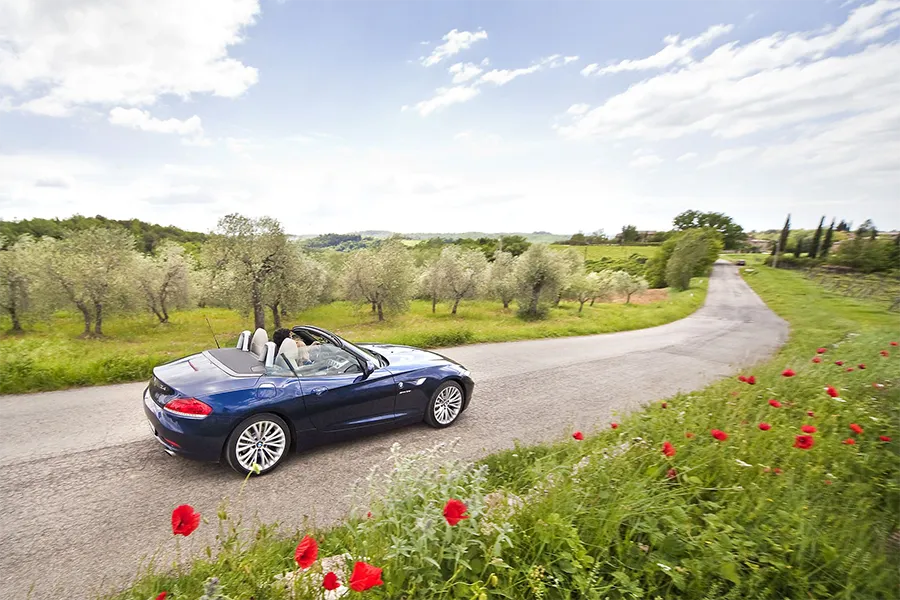 Blue convertible car driving along a scenic road between olive trees and wildflowers in the Tuscan countryside