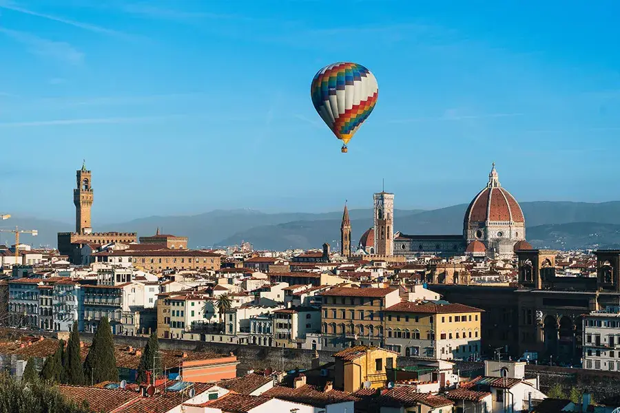 Hot air balloon flying over Florence, with a panoramic view of the Duomo, medieval towers, and mountains in the background under a clear blue sky.