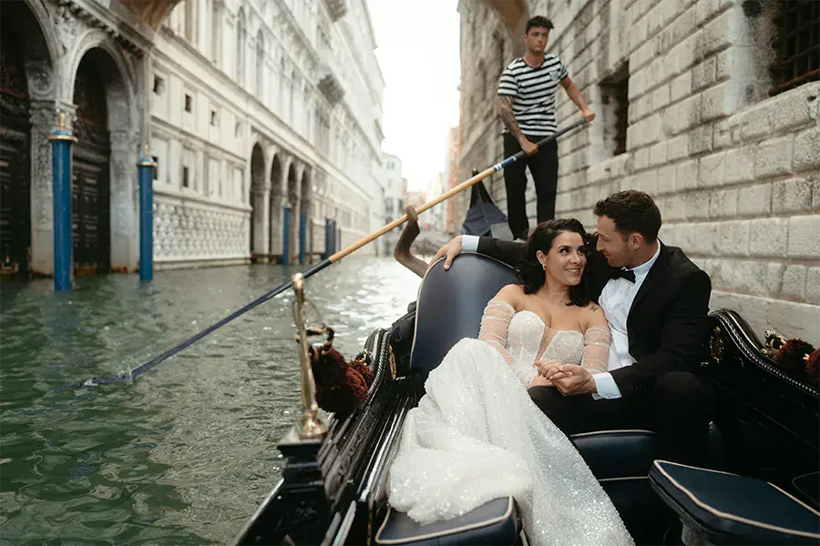 Elegant couple on a gondola ride through a Venice canal, wearing wedding attire, with a gondolier in the background, in a romantic atmosphere.