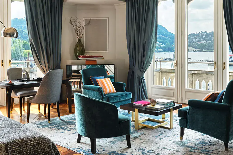 Luxurious hotel room with a view of Lake Como, featuring elegant blue-toned decor, cozy armchairs, and natural light.