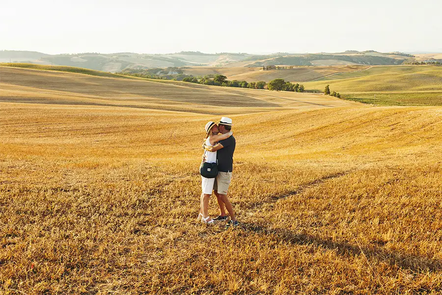 Couple hugging in the middle of a golden field in Tuscany, surrounded by rolling hills under a clear sky.