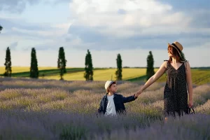 Mother and son holding hands while walking through a blooming lavender field under a partly cloudy sky.