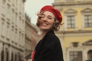 Woman smiling joyfully while wearing a red beret and black coat, standing on a charming European street.