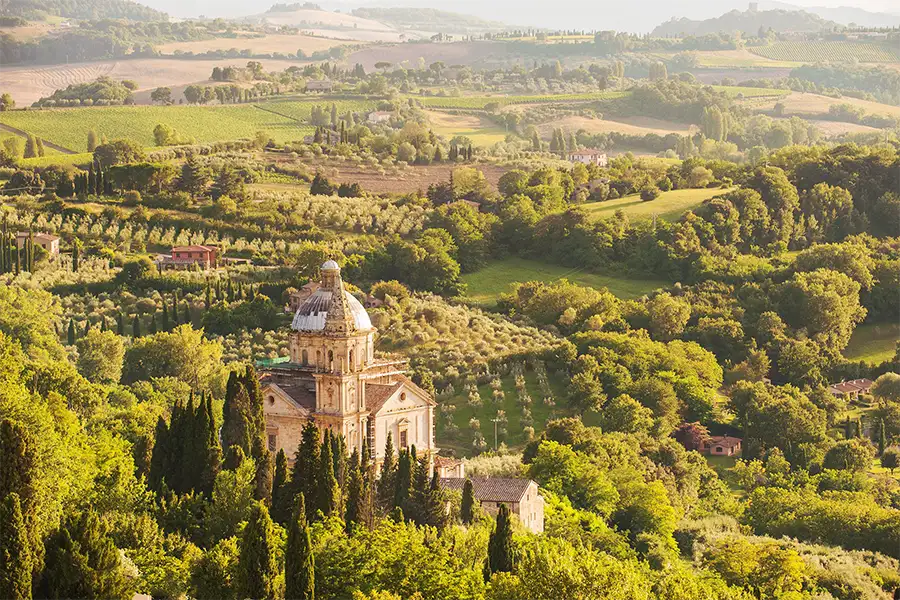 Panoramic view of Montepulciano, Tuscany, with a historic church in the foreground surrounded by lush vegetation, olive trees, and cultivated hills in the background. 