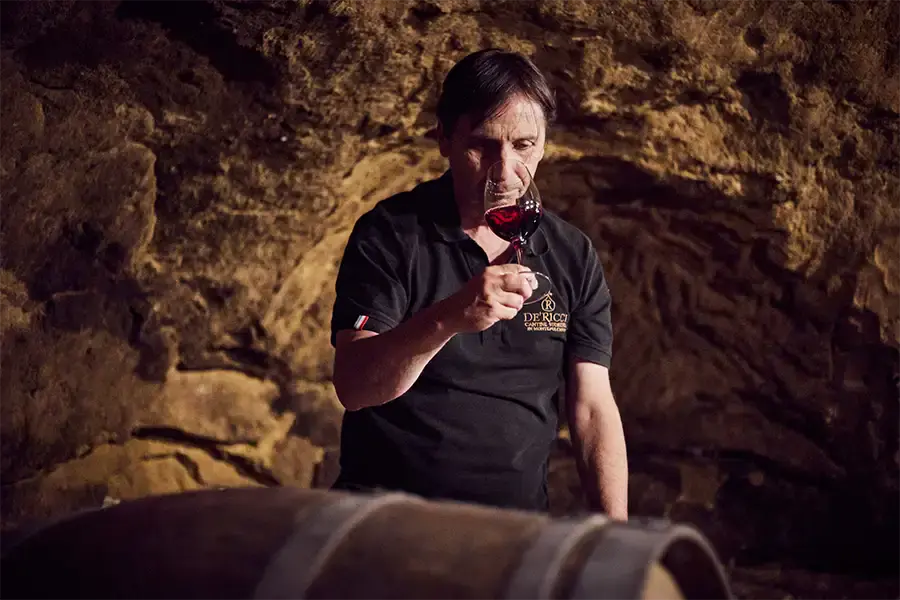 Man in a stone wine cellar examining the color of red wine in a glass beside a wooden barrel, wearing a black shirt with the De’ Ricci winery logo.