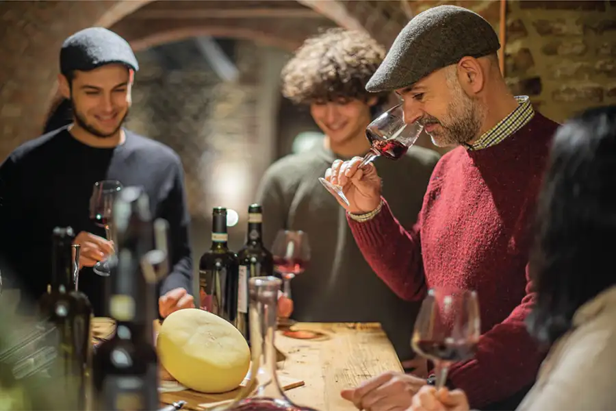 Group of people in a rustic wine cellar participating in a wine tasting, with bottles and cheese on the table; a man enjoys the aroma of red wine in his glass. 