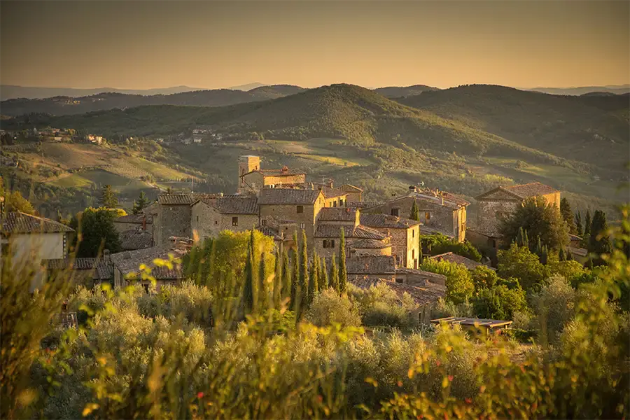 Sunset view of the medieval village of Volpaia in Tuscany, surrounded by green hills, cypress trees, and farmland, bathed in golden light. 