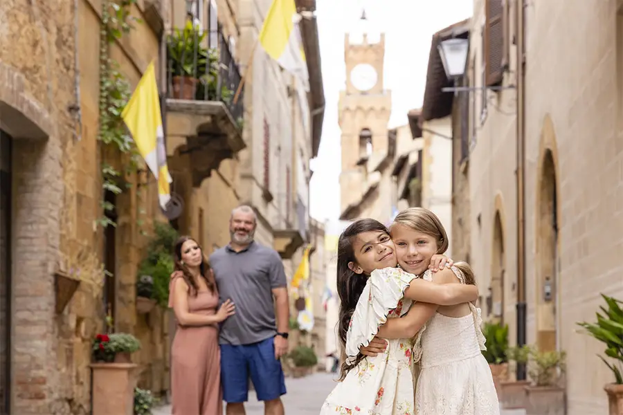 Two girls hug and smile on a charming street in a medieval town in Tuscany, with their parents in the background during a family trip to Italy.