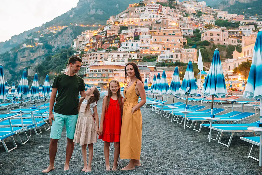Family posing on the beach in Positano, on the Amalfi Coast, surrounded by blue umbrellas and the charming colorful houses on the hillside at sunset, during a family trip to Italy.