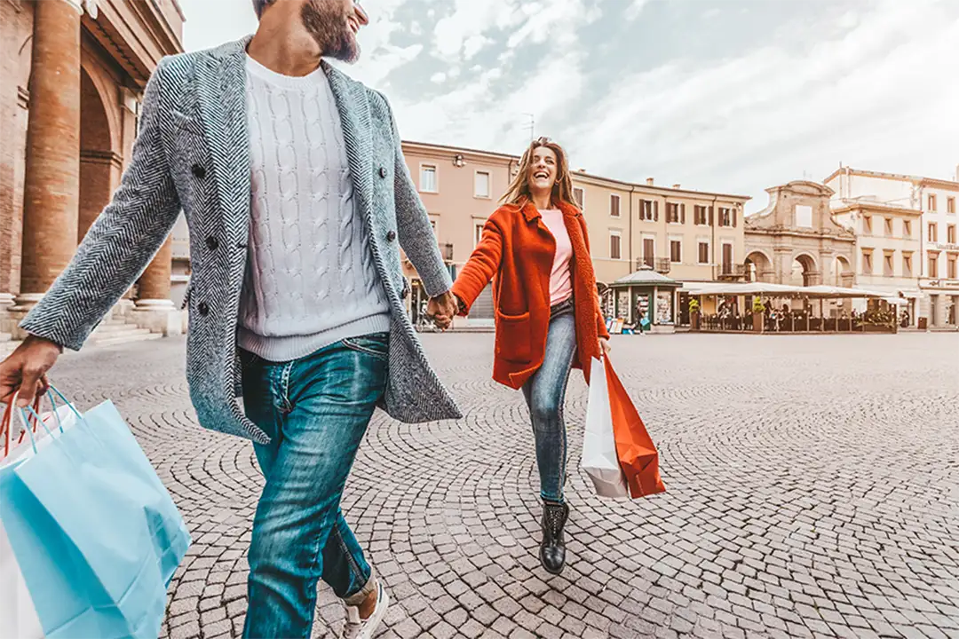 Smiling couple with shopping bags in an Italian square, embracing the casual and modern spirit of Italian fashion.
