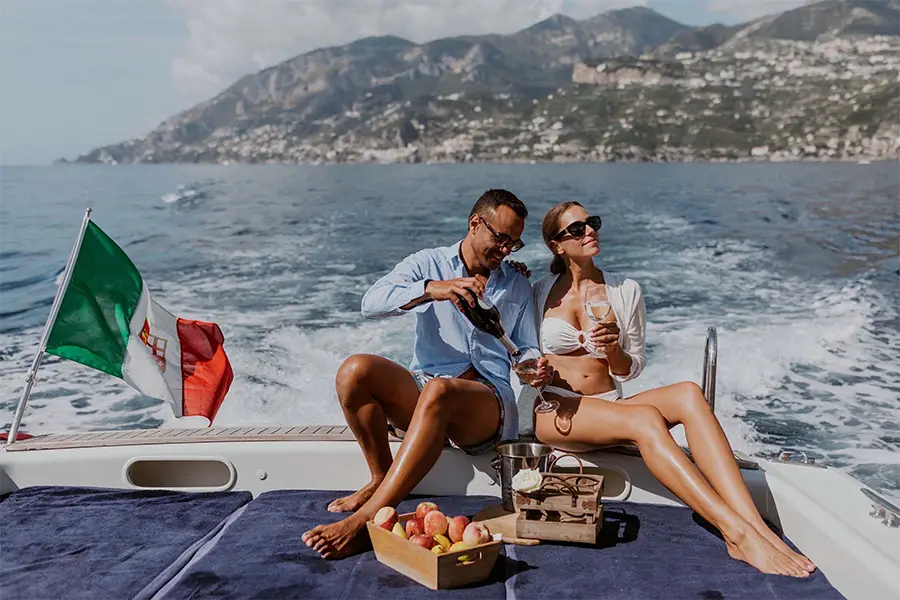 Couple enjoying a boat tour in Italy with wine glasses aboard a yacht, sailing along the Amalfi Coast with a mountainous landscape in the background and the Italian flag waving in the wind