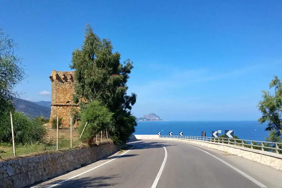 Coastal road SS113 in Sicily with sea views, stone tower, and mountains in the background