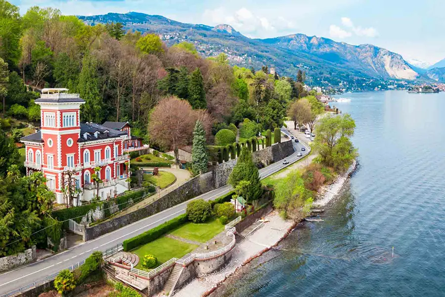 Elegant red villa on the shores of Lake Maggiore with a scenic lakeside road and mountains in the background