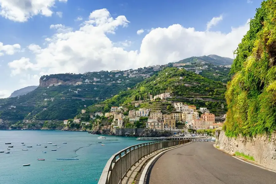 Scenic coastal road on the Amalfi Coast with hillside villages, terraced gardens, and boats floating on the blue sea