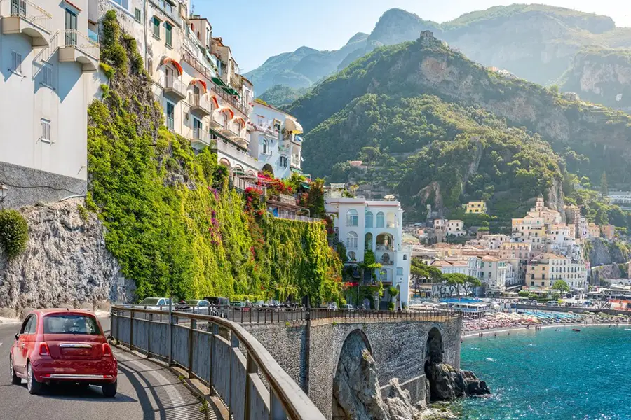 Red Fiat 500 driving along the Amalfi Coast road with colorful cliffside houses, mountains, and the Mediterranean Sea in the background